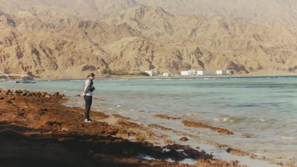 Vacker kvinna promenader ensam på stenig strand nära havet, vågorna bryts på stranden, Egypten Sinai berget på bakgrunden, slow motion, full hd — Stockvideo