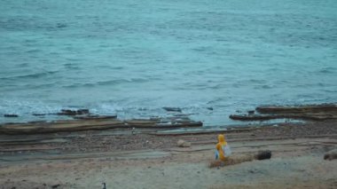 Deniz kıyısındaki taşlı sahilde tek başına oturan güzel bir kadın, dalgalar kıyıda kırılıyor, arka planda Mısır Sina dağı, yavaş çekim, tam kapasite