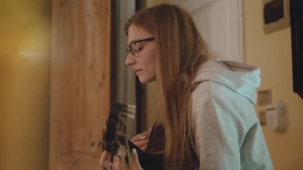Блондинка в очках играет на гитаре и поет на стуле в уютной квартире. Женщина создает музыку в помещении, замедленную, полную. — стоковое видео