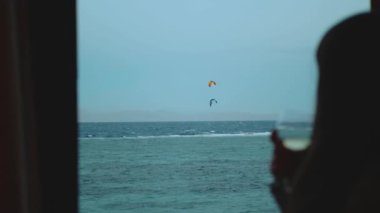 Kadın şarap içiyor ve bak insanlar Dahab Mısır 'da temiz suda nasıl sörf yapıyor. Arka planda dağlarla mavi denizi keşfetmek ve insanlar uçurtma sörfü, yavaş çekim, tam yol ileri.