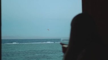 Kadın şarap içiyor ve bak insanlar Dahab Mısır 'da temiz suda nasıl sörf yapıyor. Arka planda dağlarla mavi denizi keşfetmek ve insanlar uçurtma sörfü, yavaş çekim, tam yol ileri.