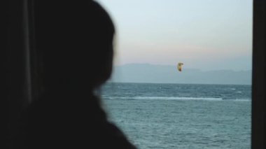 Kadın şarap içiyor ve bak insanlar Dahab Mısır 'da temiz suda nasıl sörf yapıyor. Arka planda dağlarla mavi denizi keşfetmek ve insanlar uçurtma sörfü, yavaş çekim, tam yol ileri.