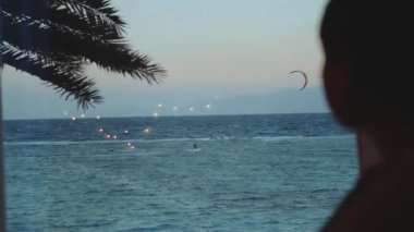 Kadın şarap içiyor ve bak insanlar Dahab Mısır 'da temiz suda nasıl sörf yapıyor. Arka planda dağlarla mavi denizi keşfetmek ve insanlar uçurtma sörfü, yavaş çekim, tam yol ileri.