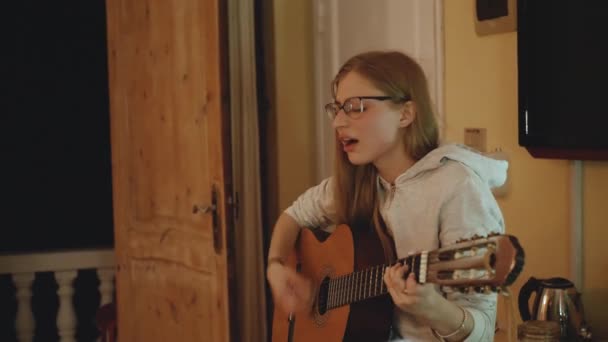Блондинка в очках играет на гитаре и поет на стуле в уютной квартире. Женщина создает музыку в помещении, 4k — стоковое видео