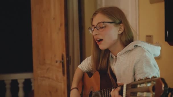 Блондинка в очках играет на гитаре и поет на стуле в уютной квартире. Женщина создает музыку в помещении, 4k — стоковое видео