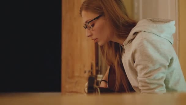 La mujer rubia en gafas toca la guitarra y canta en la silla en un acogedor apartamento. Una mujer crea música en el interior, 4k — Vídeos de Stock