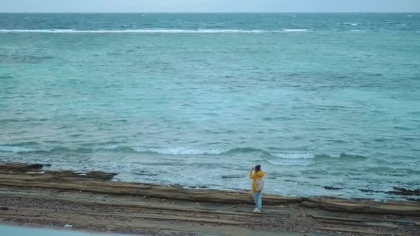 Красавица ходит и фотографирует на каменистом пляже у моря, волны ломаются на берегу, египетская гора Синай на заднем плане, 4K — стоковое видео