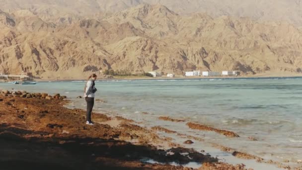 Красивая женщина, идущая одна по каменистому пляжу у моря, волны ломаются на берегу, египетская гора Синай на заднем плане, 4к — стоковое видео