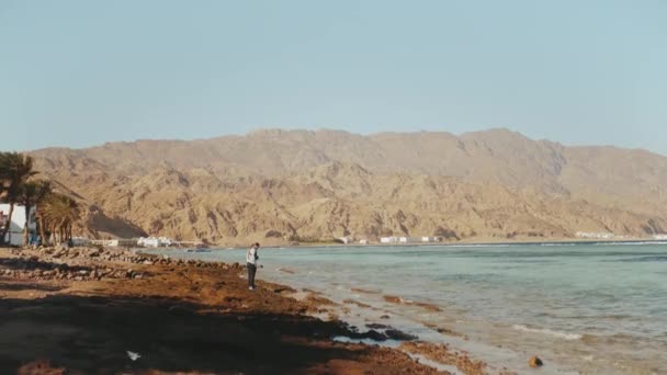 Mulher bonita andando sozinha na praia da costa pedregosa perto do mar, as ondas estão quebrando na costa, Egito Sinai montanha no fundo, 4k — Vídeo de Stock