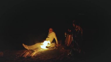 Bir grup arkadaş, gece deniz kenarında kamp ateşinin yanında eğleniyorlar.