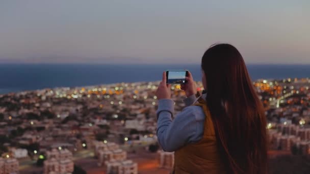 Mujer tomar una foto del mar por teléfono inteligente en la noche. Mujer permanecer en la cima de la montaña y mirando en Horizonte, 4k — Vídeo de stock