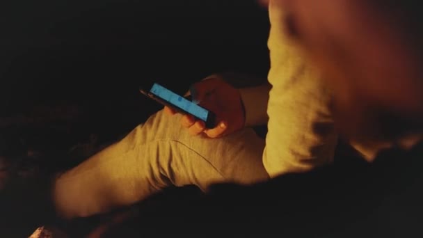 Grupo de amigos divirtiéndose cerca de la fogata en la playa cerca del mar por la noche, hombre sosteniendo teléfono inteligente, 4k — Vídeo de stock