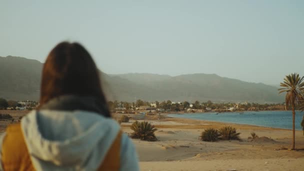 Utsikt over sandstrand og blått hav fra bak hunnen. Jente blir på stranden og leter etter Into Horizon, 4k. – stockvideo