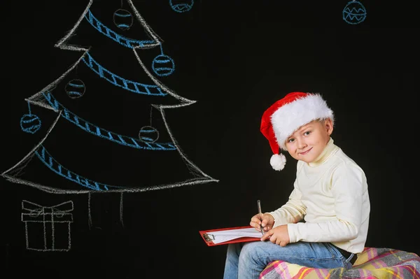 Mały chłopiec dziecko w Santa hat pisze list do Świętego Mikołaja, na ciemnym tle. W Wigilię Bożego Narodzenia — Zdjęcie stockowe