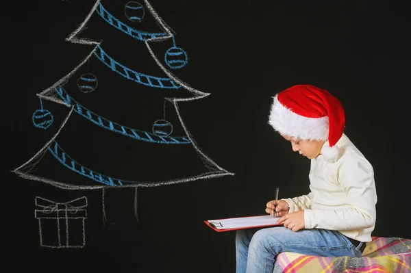 Menino bebê em chapéu de Papai Noel escreve carta para Papai Noel, em um fundo escuro. Na véspera de Natal — Fotografia de Stock