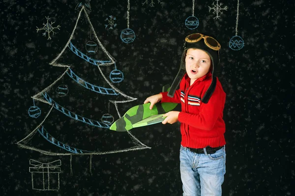 A handsome boy with a wooden toy plane , a retro helmet on a background with a painted tree — Stock Photo, Image