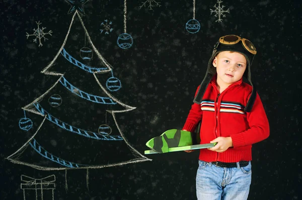 Um menino bonito com um avião de brinquedo de madeira, um capacete retro em um fundo com uma árvore pintada — Fotografia de Stock