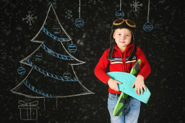 A handsome boy with a wooden toy plane , a retro helmet on a background with a painted tree — Stock Photo, Image