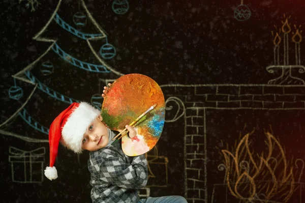 Junge mit Weihnachtsmütze auf weihnachtlichem Hintergrund, spielt mit einer Palette und Pinseln. Frohe Weihnachten — Stockfoto