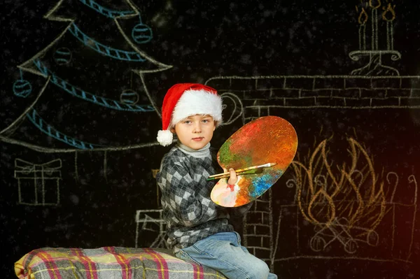 Menino em Santa chapéu no fundo de Natal, brincando com uma paleta e pincéis .Feliz Natal — Fotografia de Stock