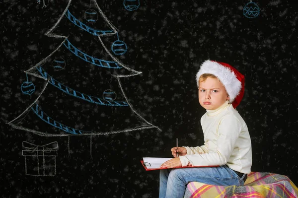 Retrato de belo menino no chapéu de Papai Noel em um fundo escuro, com árvore pintada — Fotografia de Stock