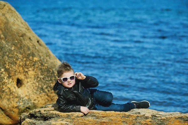 Il ragazzo con gli occhiali sul mare. Un bambino in posa in primavera sdraiato sulla pietra sullo sfondo del mare — Foto Stock