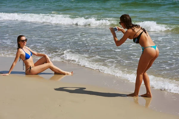 Dos mujeres jóvenes divirtiéndose en la playa, ella lleva a su novia en un teléfono móvil — Foto de Stock