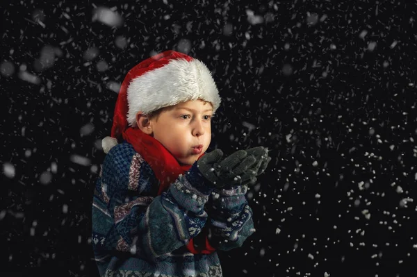 Menino feliz em Santa chapéu brinca com flocos de neve em um fundo escuro — Fotografia de Stock