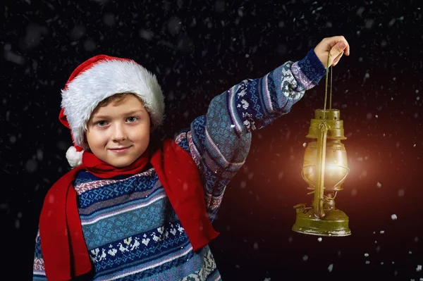 Menino de chapéu de Natal com lanterna na mão, aponta o caminho Papai Noel voando em seu trenó com a lua — Fotografia de Stock