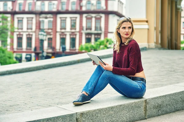 Young woman with tablet on city street . Stylish young woman in jeans and Burgundy sweater uses modern technology