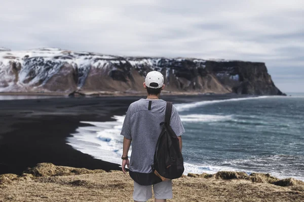 Dyrholaey, 아이슬란드에 검은 모래 해변에서 절벽에 서 있는 배낭과 젊은 남자 — 스톡 사진