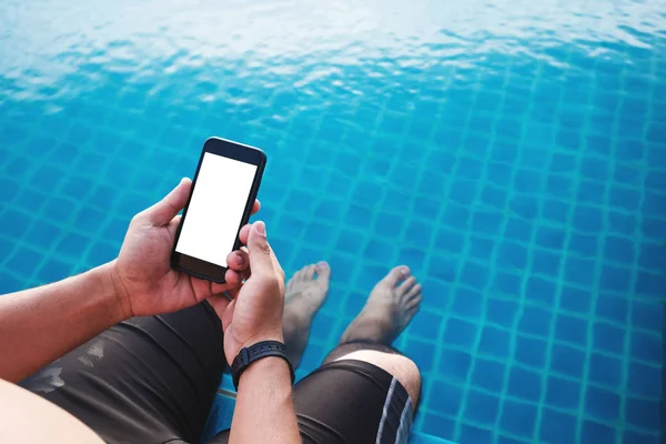 a man using mobile smart phone, blank space on screen, with legs dipping in pool in summer. Clipping path on mobile phone screen