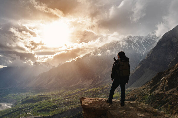 a man with backpack standing on mountain peak and bright sunlight through cloudy sky