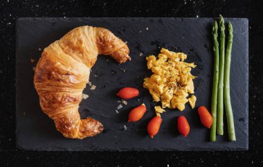 Çırpılmış yumurtalı taze kruvasan, kiraz domatesleri ve siyah taş tabakta kuşkonmaz.