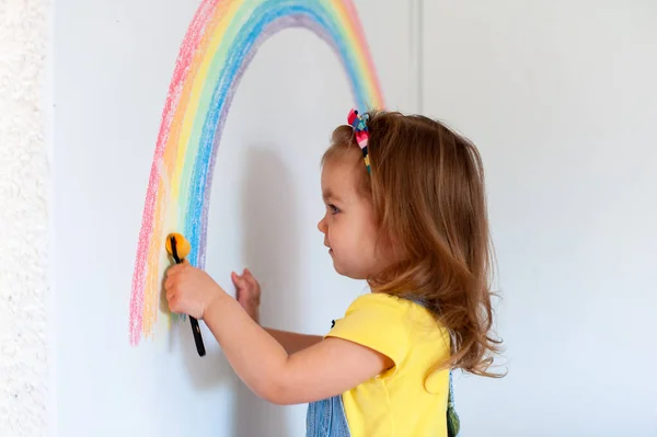 2 años de edad, chica se divierte en casa. Signo de esperanza. Coronavirus. Quédate en casa. Actividades infantiles en interiores. Chica en ropa colorida está dibujando arco iris en la pared. Chica hablando por teléfono —  Fotos de Stock