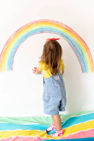 2歳の女の子は家で楽しんでいます。希望の印だ。コロナウイルス。家にいて。屋内の子供の活動。カラフルな服の女の子が壁に虹を描いている. — ストック写真