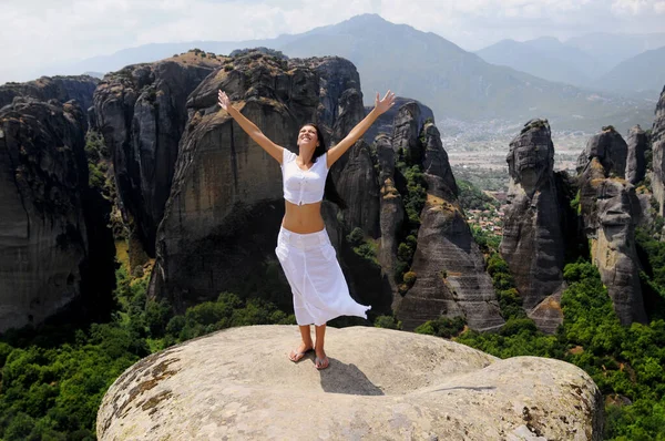 Beyaz tenli esmer kadın, elleri yukarıda, dağ manzaralı büyük bir kayanın kenarında duruyor..