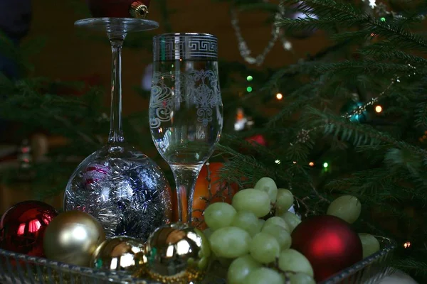 白ワインの長い脚とミサクラとクリスマスボールの中の反転ガラスを持つ1つのガラス 暗闇の中で緑のクリスマスツリーの中でトレイ上のブドウ — ストック写真