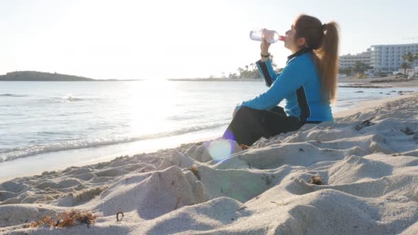 Молодая женщина в спортивной одежде питьевая вода после тренировки на пляже — стоковое видео