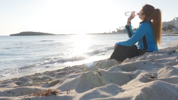 Jonge vrouw in de sport kleding drinkwater na training op het strand — Stockvideo