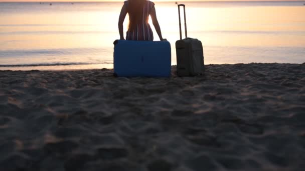 Молодая женщина сидит на чемодане на пляже — стоковое видео