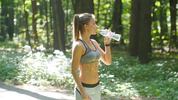 Фітнес жінка п'є воду з пляшки в сонячному лісі — стокове відео