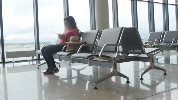 Young female passenger at the airport, using her phone while waiting for her flight — Stock Video