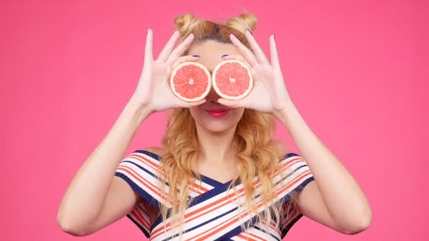 Joven mujer feliz sostiene rodajas de pomelos en sus ojos sobre fondo rosa — Vídeo de stock