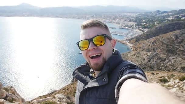 Ung man tar travel selfie på trekking utflykt dag. Mot bakgrund av havet och klipporna — Stockvideo