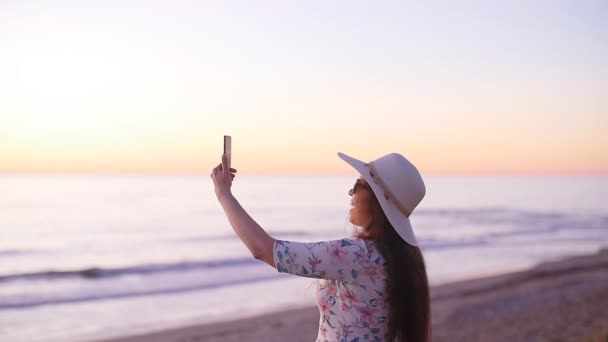 Jeune femme touristique en chapeau prenant des photos pendant le coucher du soleil sur la plage de l'océan — Video