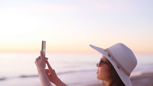 Jeune femme touristique en chapeau prenant des photos pendant le coucher du soleil sur la plage de l'océan — Video