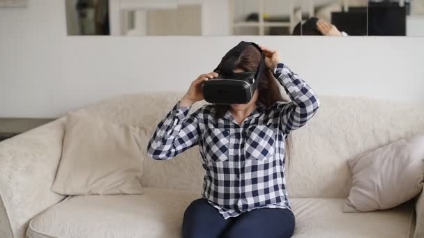 Молодая женщина в наушниках виртуальной реальности сидит дома на диване — стоковое видео