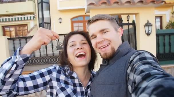 Junges Paar mit Schlüsseln steht vor dem neuen Zuhause und macht ein Selfie — Stockvideo