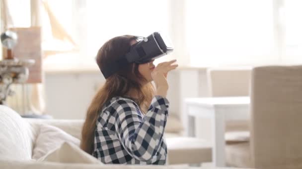 Молодая женщина с гарнитурой виртуальной реальности или 3D очки, сидящие на диване дома — стоковое видео
